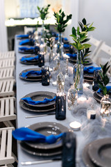 Close up of long table with porcelain crockery and shining cutlery served with green leaves and small candles. Combination of trendy blue and white colors.