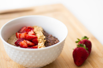 Chocolate cocoa oatmeal with almond meal, chopped strawberries and creamy peanut butter on a wooden surface. Strawberry, almonds, napkin and spoon in the background