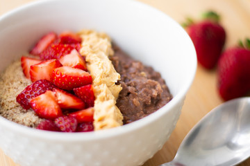 Chocolate cocoa oatmeal with almond meal, chopped strawberries and creamy peanut butter on a wooden surface. Strawberry, almonds, napkin and spoon in the background