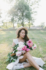 
Portrait of a young woman in a hat and an armful of peonies. Beautiful stylish girl in a summer dress, a hat and a bouquet of flowers. Girl and flowers.