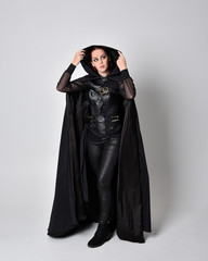 Naklejka premium fantasy portrait of a woman with red hair wearing dark leather assassin costume with long black cloak. Full length standing pose isolated against a studio background.