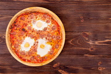 Top view of wooden table with a whole pizza with beef sausages, eggs and bacon or Brunch pizza on a round wooden platter with copy space