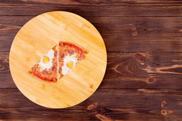 Two slice of tasty pizza with beef sausages, eggs and bacon on a wood platter which is on wooden rustic background, top view and copy space