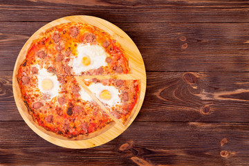 Pizza with beef sausages, eggs and bacon, with a slice removed on a round wooden platter which is on wood rustic background, top view and copy space