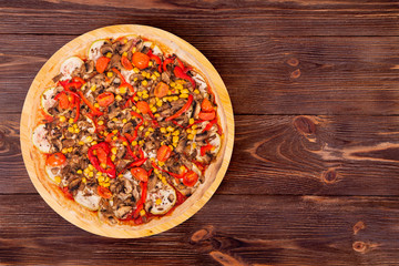 A whole delicious pizza with mushrooms, corn, cherry tomatos, courgettes and bell peppers on a round wooden plate on the wooden table, top view with copy space