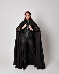 Fototapeta premium fantasy portrait of a woman with red hair wearing dark leather assassin costume with long black cloak. Full length standing pose isolated against a studio background.