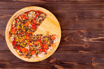 Pizza with mushrooms, corn, cherry tomatos, courgettes and bell peppers, without one slice on a wood platter which is on wooden rustic background, top view and copy space