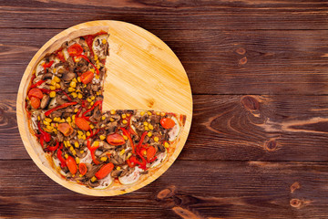 Delicious pizza with mushrooms, corn, cherry tomatos, courgettes and bell peppers, without a quarter on a round platter which is on wooden rustic background, top view and copy space