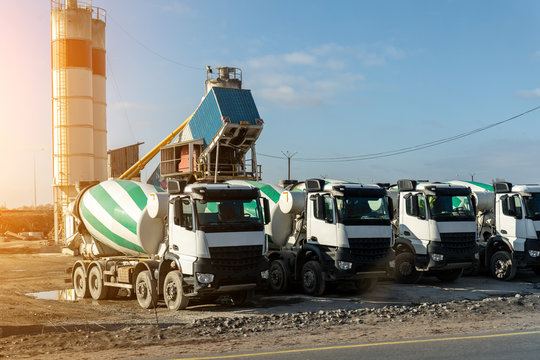 Row of many modern big mixer trucks parked against mobile temporary concrete plant factory at new asphalt road construction site morning day. Heavy machniery and industrial facilities background