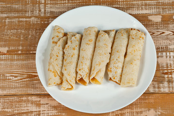 Pancakes are twisted into a tube and stuffed with cheese. Food in a white plate on a wooden table. Pancakes on a background of pink boards.