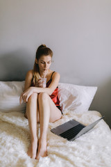 High angle of barefoot young lady in sleepwear watching film on laptop and drinking milkshake while resting on bed in weekend morning