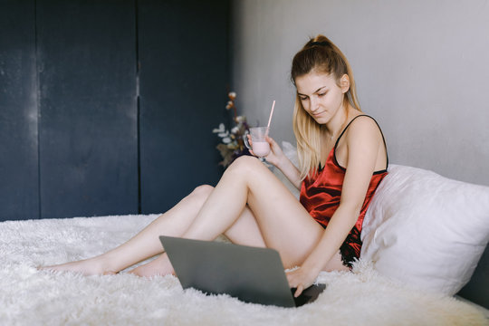Side view of young female in silk pajama enjoying milkshake and browsing laptop while sitting on bed at home