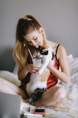 Happy young lady in pajama smiling and giving cup with milkshake to cat while sitting on bed during break in home studies