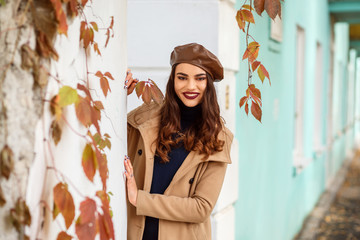 Portrait of beautiful caucasian woman spending time outdoors in autumn. Woman is dressed in dark blue sweater, beige coat and brown leather beret. Tourquoise wall in the background. 