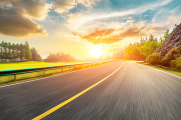 Naklejka premium Fast moving asphalt highway and green forest at sunset.