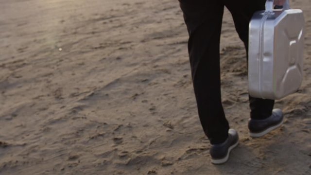 A man in a black classic suit walks along the sand and carries a metal canister in his hands.