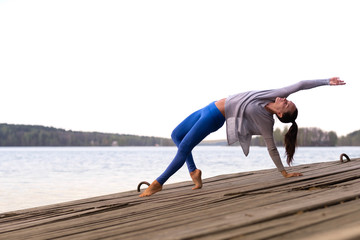 Fototapeta na wymiar A charming athletic muscular woman does yoga in blue leggings on a wooden bridge by the lake