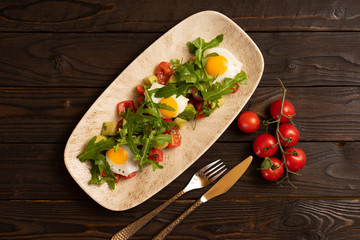 Fried eggs with cherry tomatoes, cucumber, avocado and arugula in a light plate on a dark wooden table. Healthy breakfast of eggs and vegetables, top view.