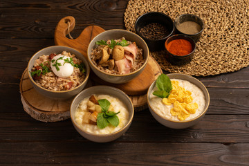 Oatmeal with cornflakes, rice porridge with fried pear, buckwheat porridge with bacon and mushrooms, and pearl barley porridge with bacon, dried tomatoes and poached egg and spices on a wooden table.