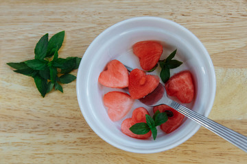 Refreshing slices of red, ripe and juicy heart-shaped watermelon in a white bowl with a fork and mint leaves on a light wooden background top view