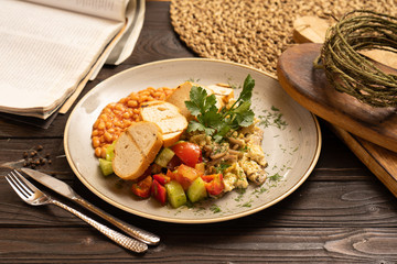 A rich breakfast of omelette beans with mushrooms, vegetables, tomatoes, bell peppers, cucumbers, parsley and dill, is beautifully served on a dark wooden table.