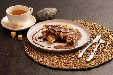 Viennese waffle with nutella, mint, and chocolate. Dessert and a cup of tea are beautifully preserved against a dark background.