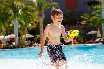 Naklejka premium Happy little kid boy jumping in the pool and having fun on family vacations in a hotel resort. Healthy child playing in water with a water gun. Laughing running child splashing