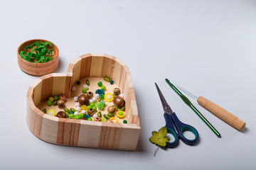 Close up of wooden heart shaped box of beads and buttons, awl, scissors and crochet for sewing and embroidery. Green and yellow set of materials for handcraft, making of bijouterie and accessories. 