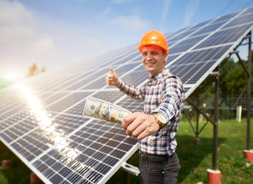 Close Up Hands Mans With Hundred Dollar Bill In It And Shows Thumb A Sign Like At The Background Of Solar Station. Business And Generation Concept. Green Ecological Power Energy Generation.