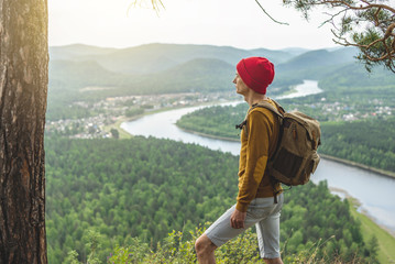 Traveler with a backpack is standing on edge of a cliff and is looking on a green valley. Freedom and unity with nature