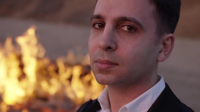 In the frame a serious handsome adult man in a classic suit and shirt looks at the camera against the background of fire in a sand quarry. the piano is burning