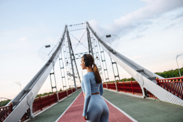Healthy active lifestyle. Pretty athlete girl in trendy sports wear with a headphones on the city bridge in a sunset light