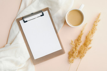 Flat lay blank paper clipboard, cup of coffee, white blanket and dry flowers on beige background. Top view home office desk table. Elegant feminine workspace concept