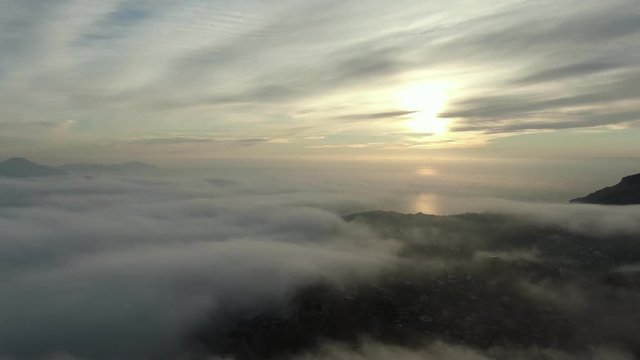 aerial survey of the sunrise over the sea covered with fog under cloudy sky and panorama of the coastal village and mountain range