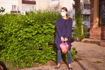 Fototapeta na wymiar A young girl in a blue jacket stands next to green shrubs in a medical mask