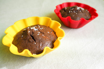 two gluten-free chocolate muffins are sprinkled with coconut shavings in a red yellow silicone mold on a beige tablecloth . sweets prepared at home