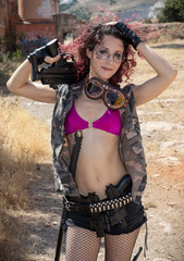 Fototapeta premium Very thin girl with pink hair and glasses dressed in military clothes carrying guns machine guns, swords and other accessories for battle in a abandoned place