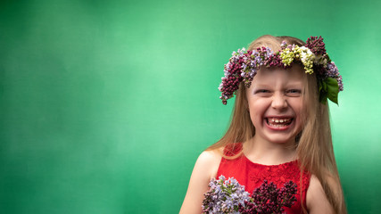 Fototapeta premium Charismatic young beautiful in a lilac wreath is having fun on a green background, with daylight. Flaunts grimace