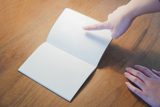 Closed up image of a notebook on a wood desk with pointing hand. Blank page.