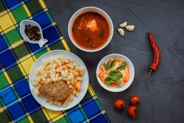 Top view od appetizing lunch set, containing meat cutlet with pasta and sauce, tomato or beetroot soup, salad made of carrot and cheese covered with parsley. Lunch served on the grey background.