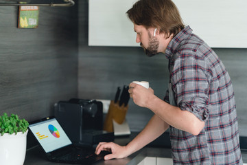 mann arbeitet im Homeoffice in der Küche am tablet, trinkt einen Kaffee und telefoniert mit bluetooth-kopfhörern