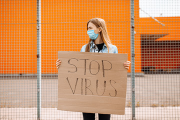 woman in a medical protective mask holds a cardboard placard with the words STOP the VIRUS, standing against a yellow wall