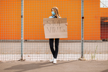 woman in a medical protective mask holds a cardboard placard with the words STOP the VIRUS, standing against a yellow wall