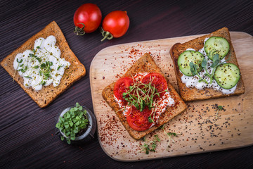 Healthy sandwiches with cream cheese, microgreens, cucumber slices and spices on cutting board with two cherry tomatoes and small jar with basil microgreen sprouts. Healthy eating, diet vegan food. 