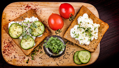 Healthy sandwiches with cream cheese, microgreens, cucumber slices and spices on cutting board with two tomatoes and small jar with basil microgreen sprouts. Healthy eating, diet vegan food. Top view