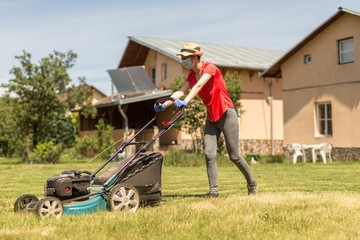 Home life during quarantine or self-isolation. A woman in her backyard mowing grass with a lawn mower on a sunny day at home wearing a surgical mask because of the coronavirus epidemic.