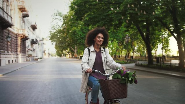 Ethnic black model in casual outfit and backpack. She smiling, riding a bicycle with lilac in basket by deserted city avenue. Slow motion