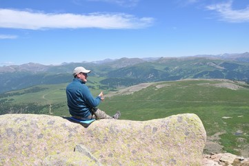 Naklejka premium A man in a sweatshirt and a baseball cap sits on a stone covered with lichen over a mountain valley