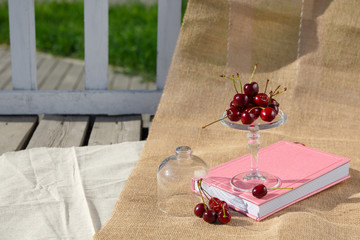Still life and food photo. Picnic in nature on a sunny day. Footed service plate and mini dome with cherry berries is on the book and on burlap, on the wooden floor from old boards