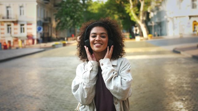 Afro-american model in headphones and casual outfit. She smiling, dancing, listening to the music while walking by deserted street of city. Close up